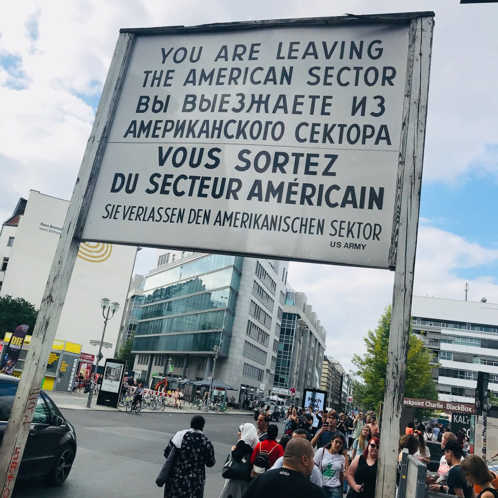 Skylt som markerar utgången från den amerikanska sektorn i Berlin, med text på engelska, ryska, franska och tyska, omgiven av en folkmassa och moderna byggnader.