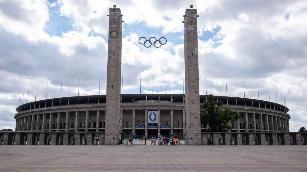 En bild av Olympiastadion i Berlin, med stora pelare och de olympiska ringarna svävande ovanför, under en molnig himmel.