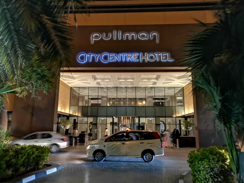 Entrén till Pullman Dubai Creek City Centre Hotel, med bilar som kör förbi och palmträd i förgrunden.