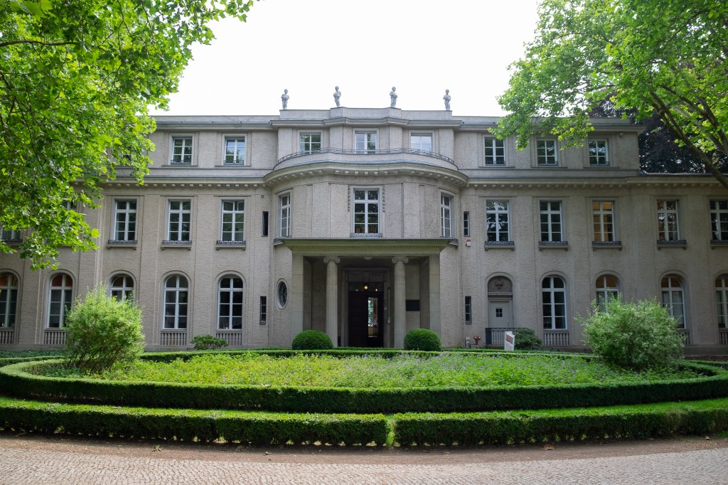 En stor och pampig villa omgiven av grönska, med en välskött trädgård framför. Byggnaden har flera fönster och en klassisk arkitektur.