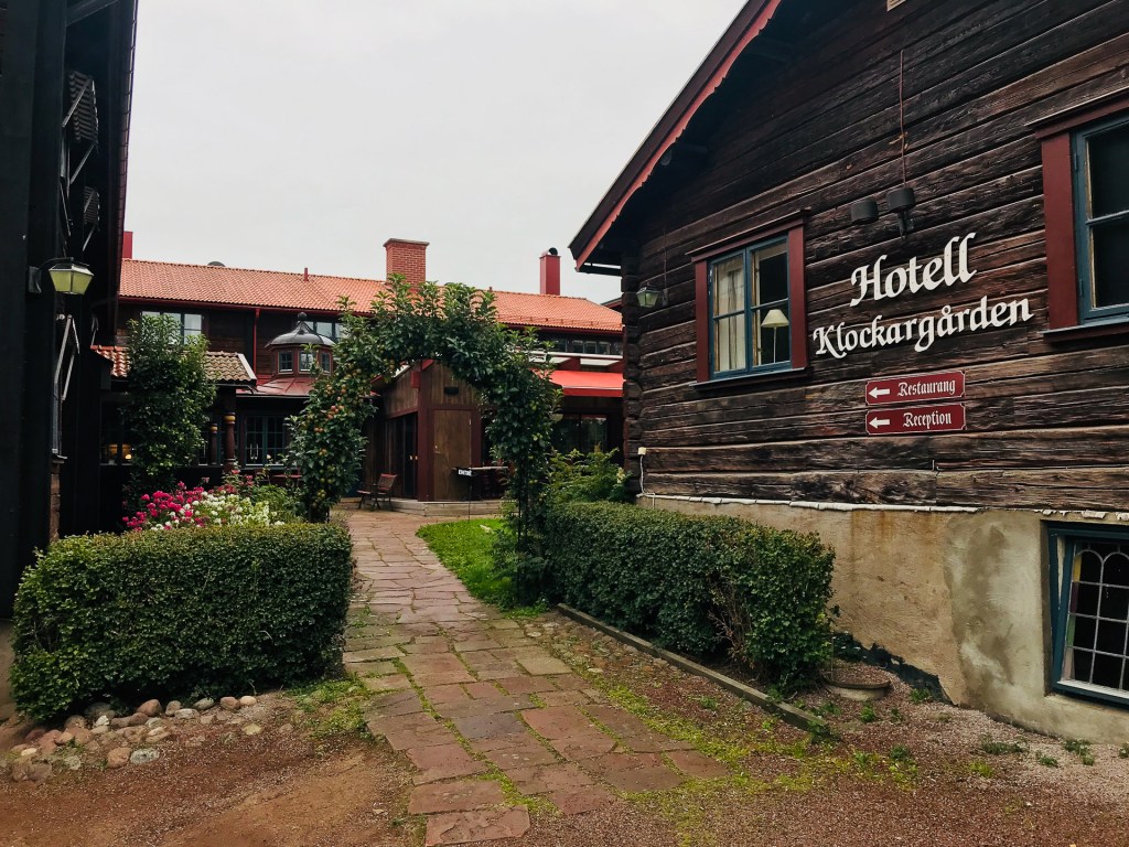 Ingången till Hotell Klockargården i Tällberg, Dalarna, med en grönskande trädgård och blomsterarrangemang.
