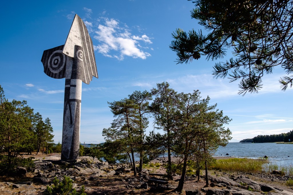 En stor, stiliserad skulptur av en fisk eller fågel i svart och vitt, omgiven av tallar och en vy av sjön i bakgrunden.