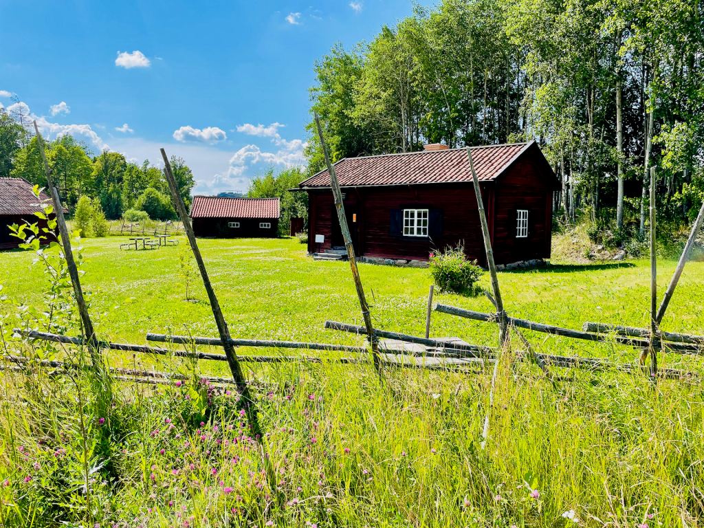 Ett soldattorp med röda väggar och tak i ett grönt fält omgiven av träd, med en staket av grenar i förgrunden.