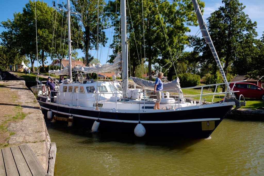 En segelbåt ligger i en sluss med en person som står på däcket. Grönska och hus syns i bakgrunden.