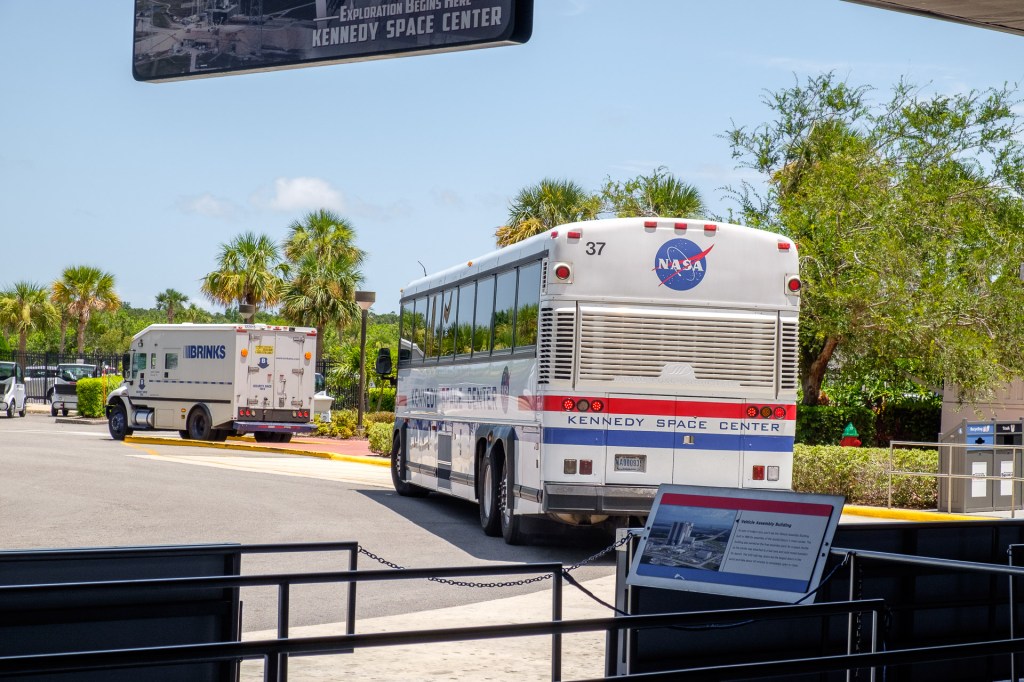 En Kennedy Space Center-buss parkerad vid ingången, med palmträd och en säkerhetsbil i bakgrunden.