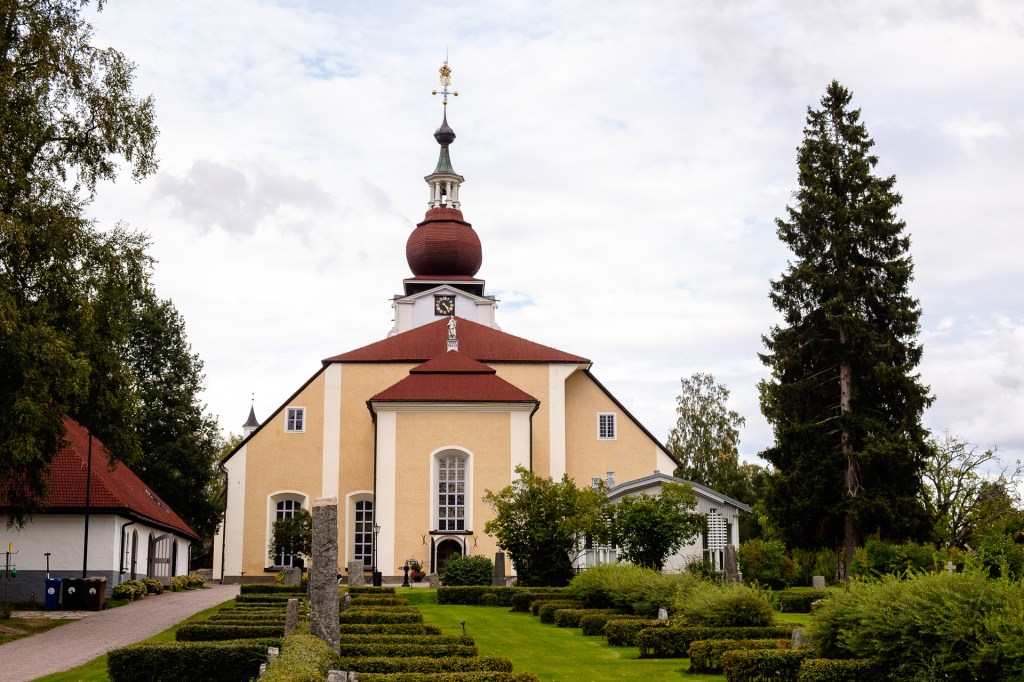 En gul kyrka med röd takspira omgiven av grönområden och träd, med en mörk himmel i bakgrunden.
