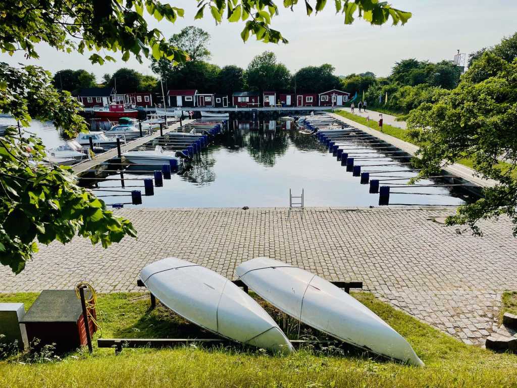 Utsikt över en hamn med båtar förtöjda vid kajen och vita segelbåtar i förgrunden, omgiven av grönska och byggnader i bakgrunden.