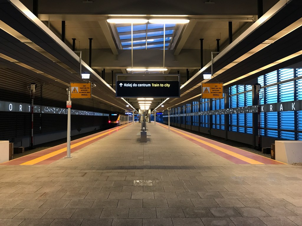 En station med plattformar och skyltar för tåg till Kraków. Det är en modern miljö med blå ljusinslag och ett tak som släpper in naturligt ljus.