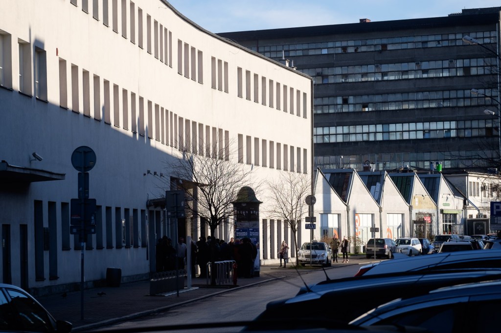 En gata i Kraków med moderna och äldre byggnader, inklusive ett flerfamiljshus och en taxistation. Människor står och väntar på sidan av vägen, medan bilar parkerar längs kanten.