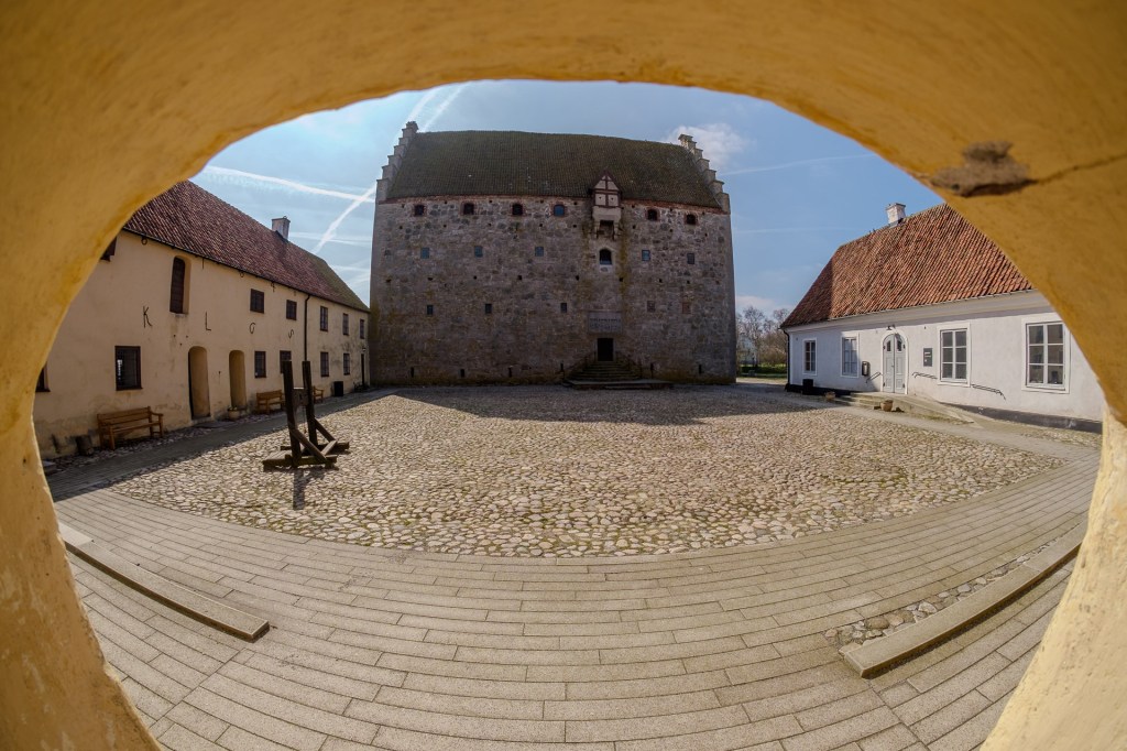 Utsikt över Glimmingehus borg genom en rundad öppning, med den medeltida borgen synlig i bakgrunden omgiven av stenläggning och byggnader.