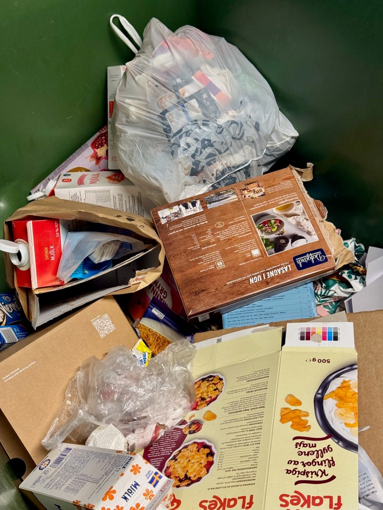 En överfull soptunna med skräp som innehåller plastpåsar, kartonger och annat avfall, inklusive pizzakartonger och matförpackningar.