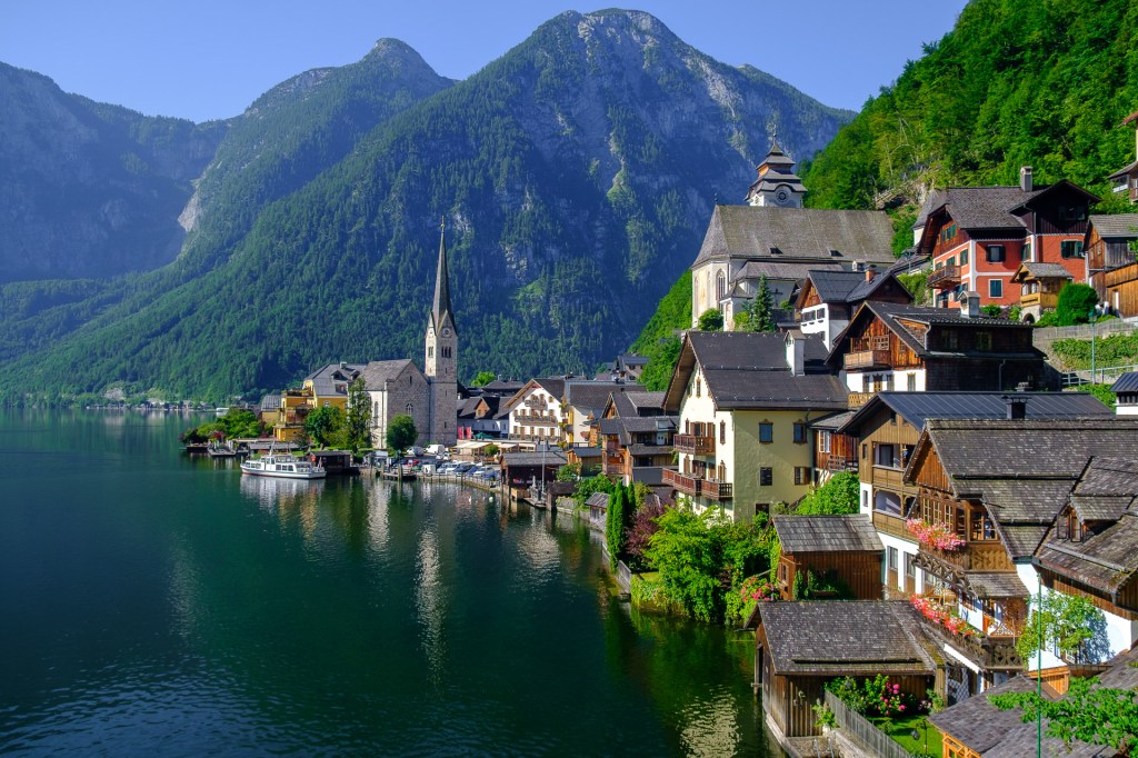 En panoramabild av den pittoreska byn Hallstatt i Österrike, med traditionella alpstugor, en kyrka och den spegelblanka sjön Hallstätter See, omgiven av mäktiga berg.