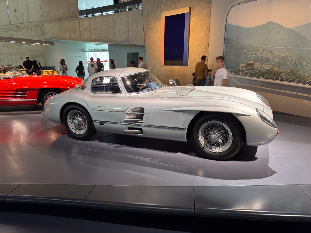 En klassisk Mercedes-bil, 300 SLR Uhlenhaut Coupé, med måsvingedörrar och elegant design, står i ett modernt museum omgiven av besökare. I bakgrunden syns en andra röd bil och en vägg med landskapsbild.