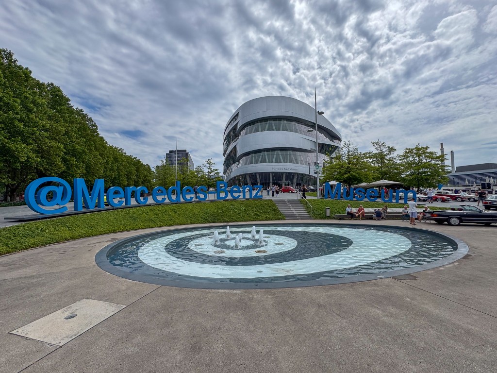 Entrén till Mercedes-Benz Museum med stora blå bokstäver som visar museets namn och en modern byggnad i bakgrunden.