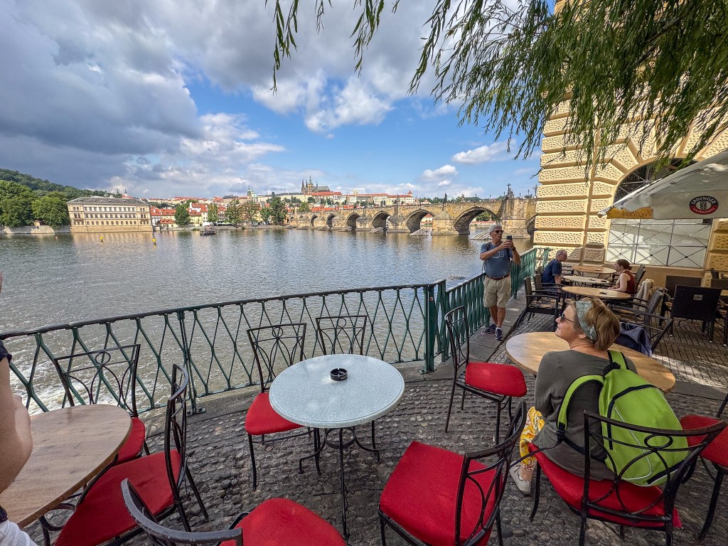 Utsikt över Vltava-floden med Karlsbron i bakgrunden, sittplatser vid vattnet och besökare som njuter av atmosfären.