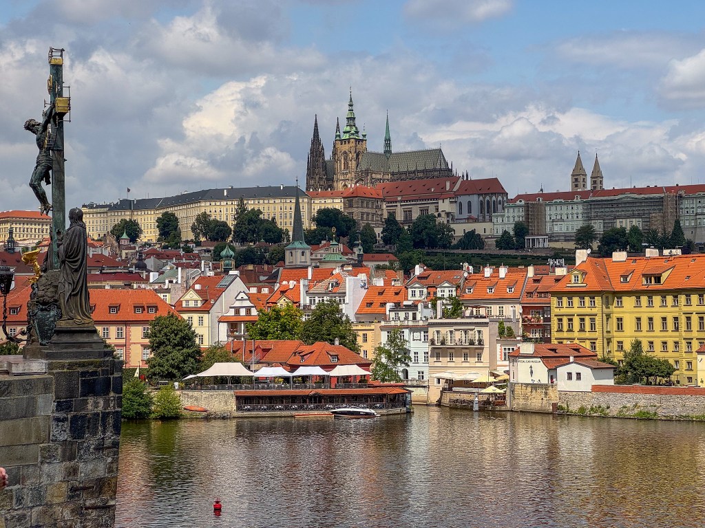 Utsikt över Prag med Prags slott synligt i bakgrunden, omgiven av historiska byggnader med röda tak och en flod i förgrunden.