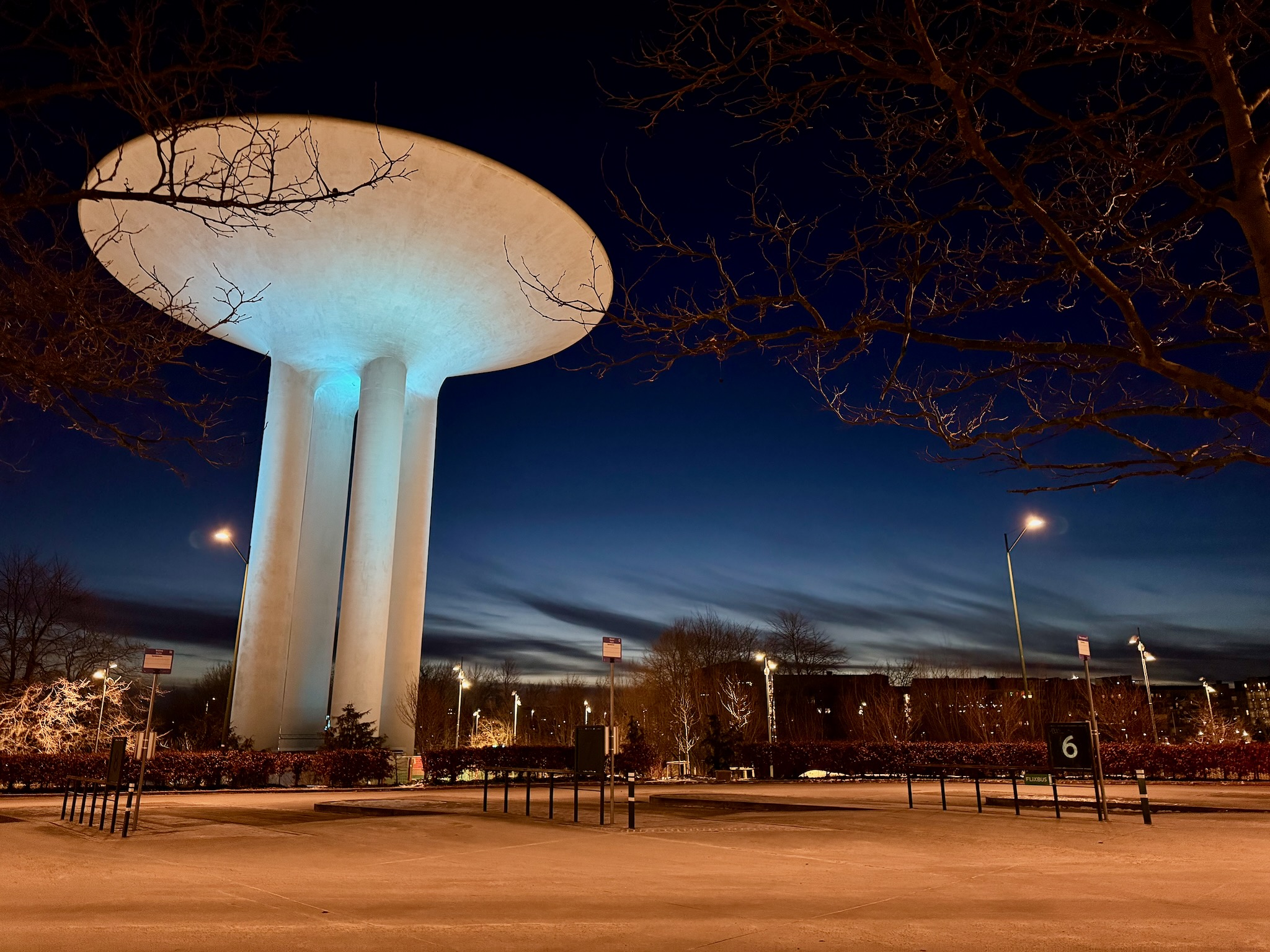 En vattentorn med stora pelare, belyst av blått ljus, mot en skymningshimlen. Trädgrenarna i förgrunden och en parkeringssituation synlig i bilden.