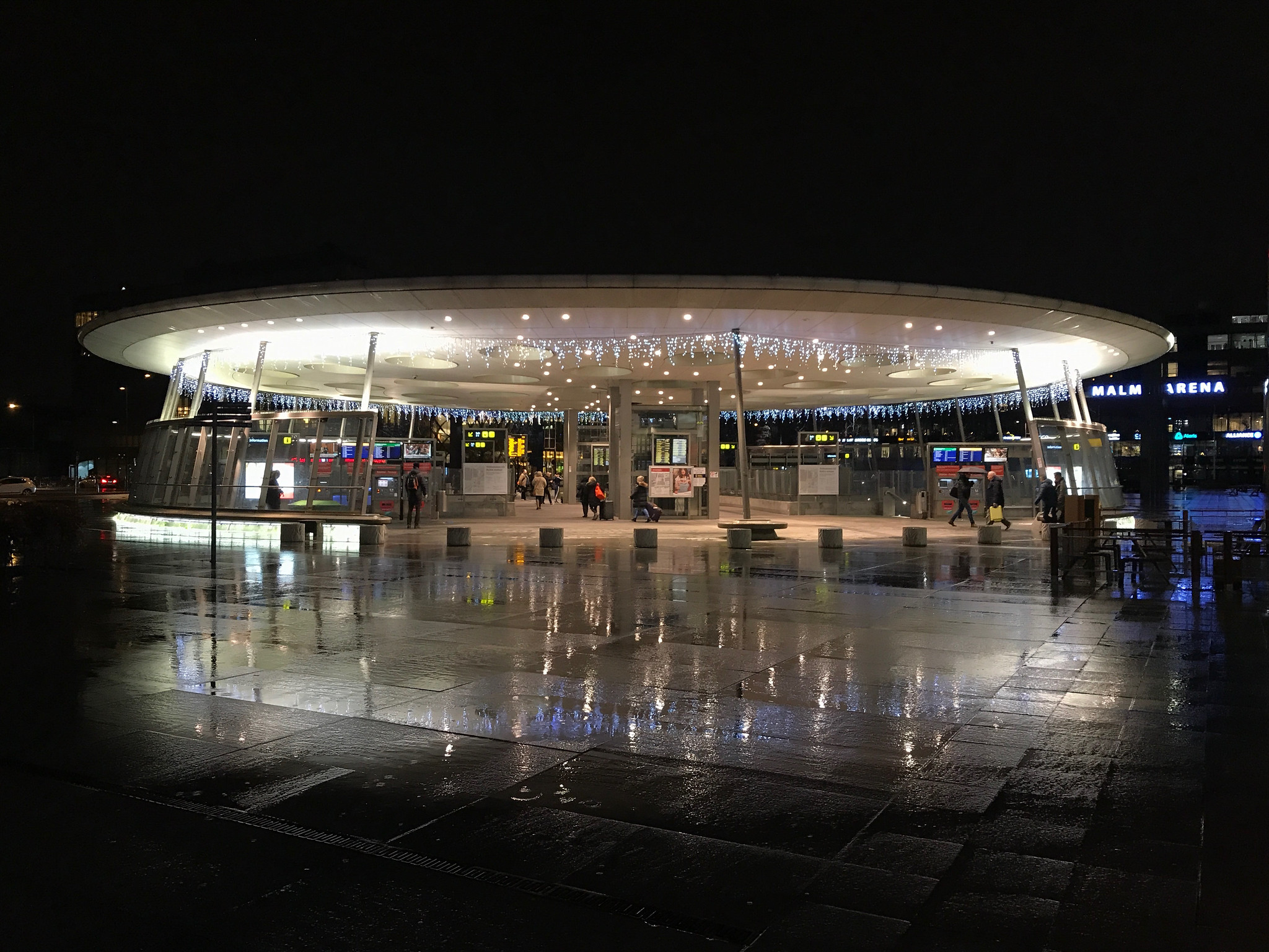 En modern tågstation i Hyllie med belysning och reflektioner på marken, fotograferad på kvällen.